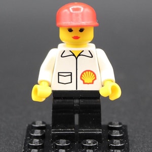 May include: A Lego minifigure of a person wearing a red hard hat and a white jacket with the Shell logo. The figure has black pants, a yellow head, and is standing on a black base.