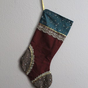 May include: A Christmas stocking with a burgundy, teal, and brown fabric design. The stocking has gold trim and lace accents.