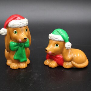 May include: Two ceramic dachshund figurines, one upright and one lying down. Both dogs are brown with green or red bows, and wear Santa hats with white trim. The figurines have black eyes and pink noses.