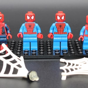 May include: Four Spider-Man Lego minifigures with red and blue suits, on black Lego baseplates. Two white spider webs are in the foreground. The figures have red masks and various suit designs.