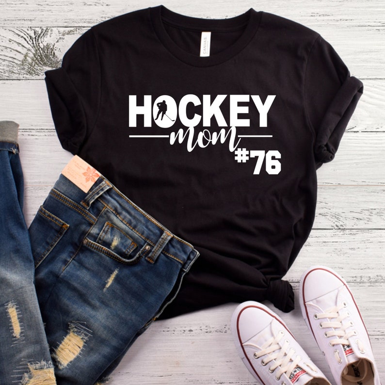 May include: Black t-shirt with white text that reads "HOCKEY mom #76". A hockey player silhouette is above the word "mom". The shirt is displayed on a white wood background with a pair of white sneakers and blue jeans.