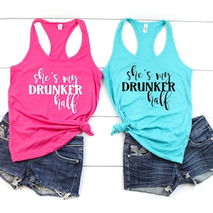May include: Two tank tops with the text "She's my Drunker half" in black and white lettering. The tank tops are pink and turquoise blue and are tied at the front. The tank tops are laying on a white surface with denim shorts.