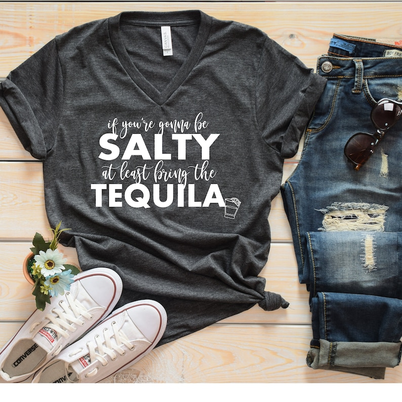 Puede incluir: Una camiseta gris oscuro con cuello en V con el texto "if you're gonna be SALTY at least bring the TEQUILA." La camiseta se combina con vaqueros azules, zapatillas blancas y gafas de sol sobre una superficie de madera.