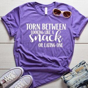 May include: Purple t-shirt with white text that reads "Torn between looking like a snack or eating one".