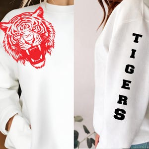 May include: White sweatshirt featuring a large red tiger graphic on the front. The sleeve displays the word "TIGERS" in black block letters. The sweatshirt has a crew neck and long sleeves.