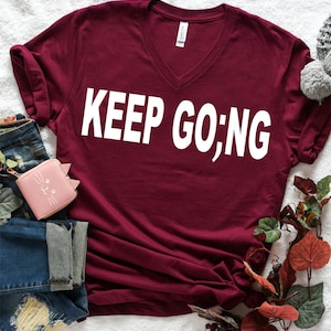 May include: Burgundy v-neck t-shirt with the text "KEEP GOING" in white lettering.