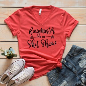 May include: Red V-neck t-shirt with the text "Ringmaster of the Shit Show" in black lettering. The text is styled with a circus tent graphic on either side of the text.