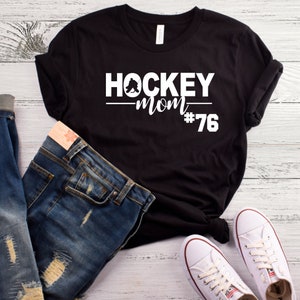 May include: Black t-shirt with white text that reads "HOCKEY MOM #76". A hockey player silhouette is above the word "MOM". The shirt is laid out on a woodgrain surface with a pair of blue jeans and white sneakers.