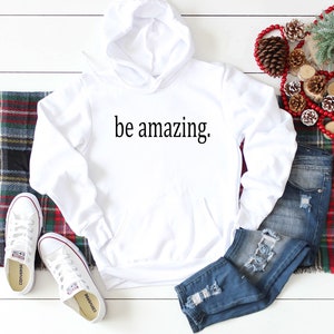 May include: White hooded sweatshirt with the text "be amazing." printed on the front. The sweatshirt is laid out on a white surface with a pair of white sneakers, blue jeans, and red and green plaid fabric.