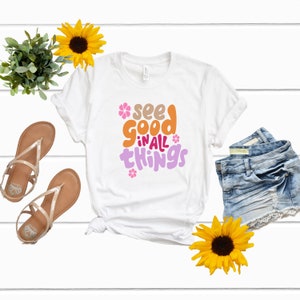Op de afbeelding: Wit T-shirt met de tekst "see good in all things" in kleurrijke, gestileerde letters met roze bloemen.