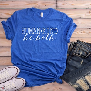 May include: A blue t-shirt with the words "HUMAN KIND be both" in white script. The shirt is paired with blue jeans and white sneakers on a wooden background. Casual wear.