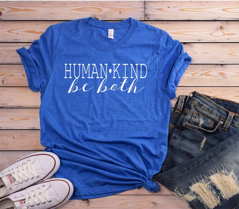 May include: A blue t-shirt with the words "HUMAN KIND be both" in white script. The shirt is paired with blue jeans and white sneakers on a wooden background. Casual wear.