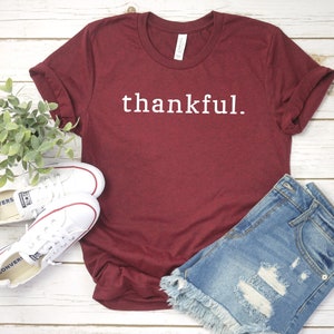 May include: A maroon t-shirt with the word "thankful." printed in white on the front. The shirt is laying on a white wooden surface with a pair of white sneakers and a pair of blue denim shorts.