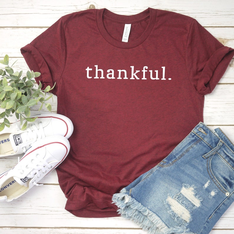 May include: A maroon t-shirt with the word "thankful." printed in white on the front. The shirt is laying on a white wooden surface with a pair of white sneakers and a pair of blue denim shorts.