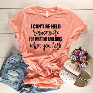 Könnte beinhalten: Ein pfirsichfarbenes T-Shirt mit dem Text "Ich kann nicht für das verantwortlich gemacht werden, was mein Gesicht tut, wenn du sprichst" in Schwarz gedruckt.