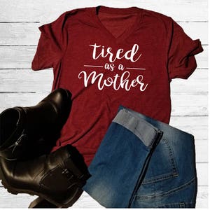 May include: A maroon v-neck t-shirt with the text "tired as a mother" in white cursive lettering.