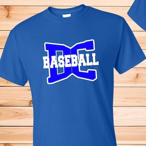 May include: A royal blue short-sleeved t-shirt featuring a large graphic on the chest. The graphic consists of a bold, stylized blue letter D and C with a white outline, overlaid with the word BASEBALL in white capital block letters.