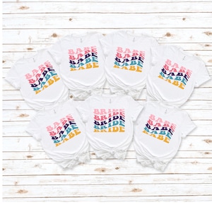 May include: Six white t-shirts with a retro design. Five shirts say "Babe" in a rainbow of colors. One shirt says "Bride" in a rainbow of colors.