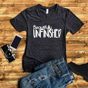 May include: A dark gray V-neck t-shirt with the text "Beautifully Unfinished" in white lettering. The shirt is shown on a wooden background with a pair of blue jeans and a camera.