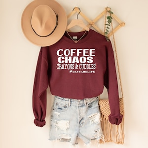 Op de afbeelding: Bordeauxkleurige trui met witte tekst die "Coffee Chaos Crayons & Cuddles #DaycareLife" luidt.