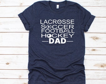 Puede incluir: Camiseta azul marino con texto blanco: "LACROSSE SOCCER FOOTBALL HOCKEY DAD" e iconos deportivos. La camiseta es de material suave y tiene mangas enrolladas.