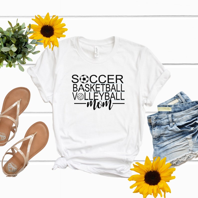 May include: White t-shirt with black text that reads "Soccer Basketball Volleyball Mom" with a soccer ball, basketball, and volleyball graphic.