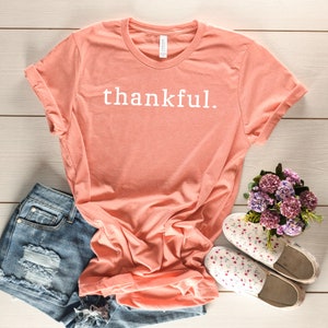 May include: A coral colored t-shirt with the word "thankful" printed in white on the front. The shirt is laid flat on a white wooden surface with a pair of floral print canvas shoes and a pair of denim shorts.