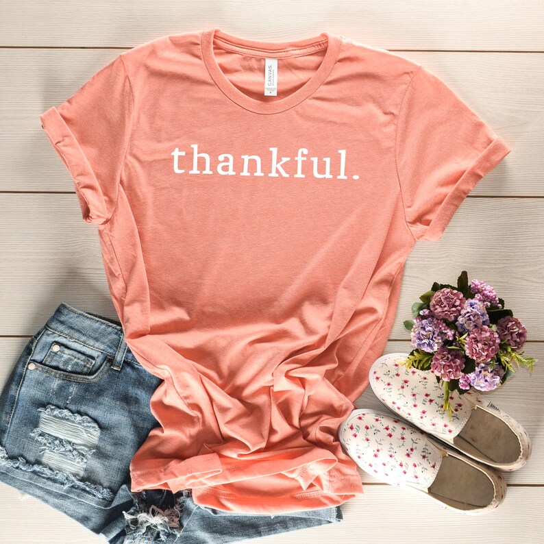 May include: A coral colored t-shirt with the word "thankful" printed in white on the front. The shirt is laid flat on a white wooden surface with a pair of floral print canvas shoes and a pair of denim shorts.