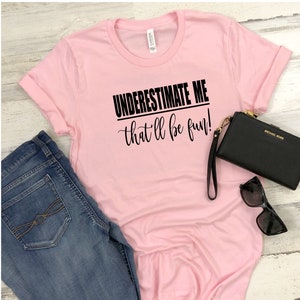 Könnte beinhalten: Rosa T-Shirt mit dem Aufdruck "UNDERESTIMATE ME that'll be fun!" in Schwarz.