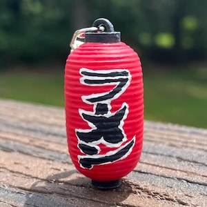 May include: A red Japanese lantern keychain with black Japanese characters. The lantern is cylindrical with a black top and bottom. The keychain is on a wooden surface with a blurred green background.