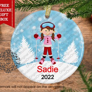 May include: A round Christmas ornament with a blue background and white snowflakes. A cartoon girl wearing a pink jacket, red pants, and a ski mask is skiing down a snowy hill. The ornament has the name "Sadie" and the year "2022" written on it.
