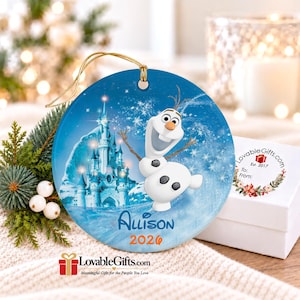 May include: A round blue ceramic ornament featuring a white snowman character with a carrot nose and twig arms, a blue castle, and the text "Allison" and "2026" printed in blue and orange. The ornament includes a gold hanging string.