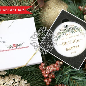 May include: White ceramic Christmas ornament with a gold leafy design and the text "Our 1st Christmas as Mr & Mrs SMITH 2022". The ornament is in a white gift box with a gold ribbon.