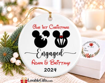 May include: A round white ceramic ornament with a gold hanging string. The design features two black mouse-ear silhouettes, one with a white bow and one with a tuxedo, with the text Our 1st Christmas Engaged, Adam & Brittany, and 2024.
