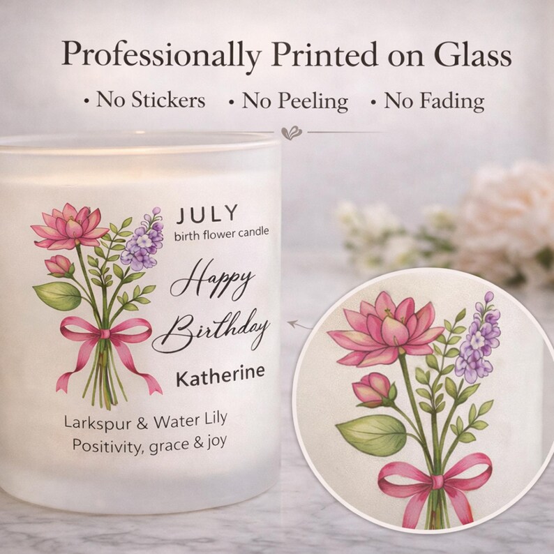 May include: A white glass candle with a floral design and the text "Happy Birthday Katherine." The design includes pink lotus flowers, purple larkspur, and a pink ribbon. The candle is labeled "July birth flower candle."