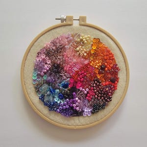 May include: A colorful beaded embroidery art piece in a wooden hoop. The design features a circular pattern of beads in a rainbow of colors, including pink, orange, purple, and blue. The beads are of various sizes and shapes, creating a textured effect.