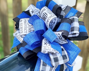 May include: A blue and black ribbon bow with the text "Blue Line, Protect & Serve, Hero & Strong, Enforce & Serve" on it. The bow is tied around a white post.