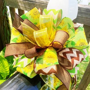 May include: A decorative bow featuring layers of yellow, brown, and green ribbons. The bow includes ribbons with sunflower patterns, transparent yellow accents, and a brown chevron design. The bow is tied around a dark object.