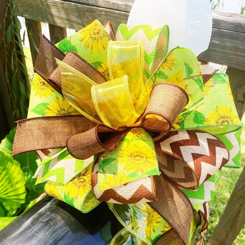 May include: A decorative bow featuring layers of yellow, brown, and green ribbons. The bow includes ribbons with sunflower patterns, transparent yellow accents, and a brown chevron design. The bow is tied around a dark object.