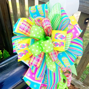 May include: A colorful mailbox bow with a summer theme. The bow is made of multiple ribbons with different patterns, including flip flops, polka dots, stripes, and plaid. The colors are bright and cheerful, including pink, yellow, green, blue, and white.