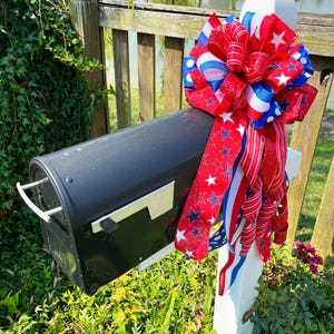 May include: A black mailbox with a white post is decorated with a large red, white, and blue bow. The bow features ribbons with stars and stripes, creating a patriotic theme. The mailbox is surrounded by green foliage.