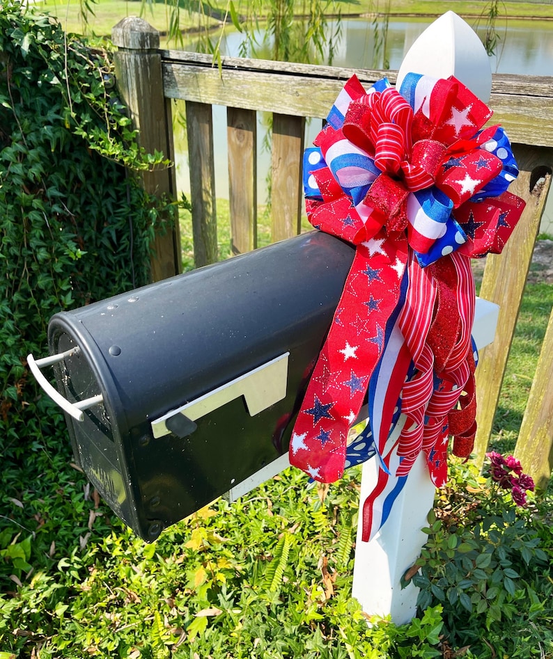 May include: A black mailbox with a white post is decorated with a large red, white, and blue bow. The bow features ribbons with stars and stripes, creating a patriotic theme. The mailbox is surrounded by green foliage.