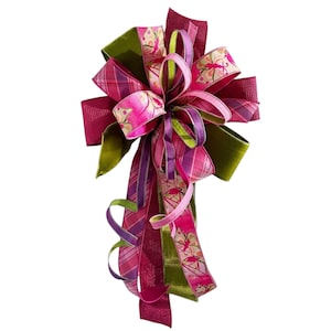 May include: A decorative bow featuring multiple ribbons in shades of magenta, olive green, and light pink. The design includes a mix of solid textures, plaid patterns, and a butterfly print. Several long, curled ribbon tails hang downward.