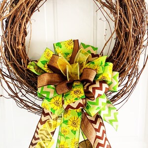May include: A grapevine wreath adorned with a large bow. The bow features ribbons with sunflower patterns, brown chevron stripes, and solid brown and yellow accents. The wreath is a decorative item for doors or walls.
