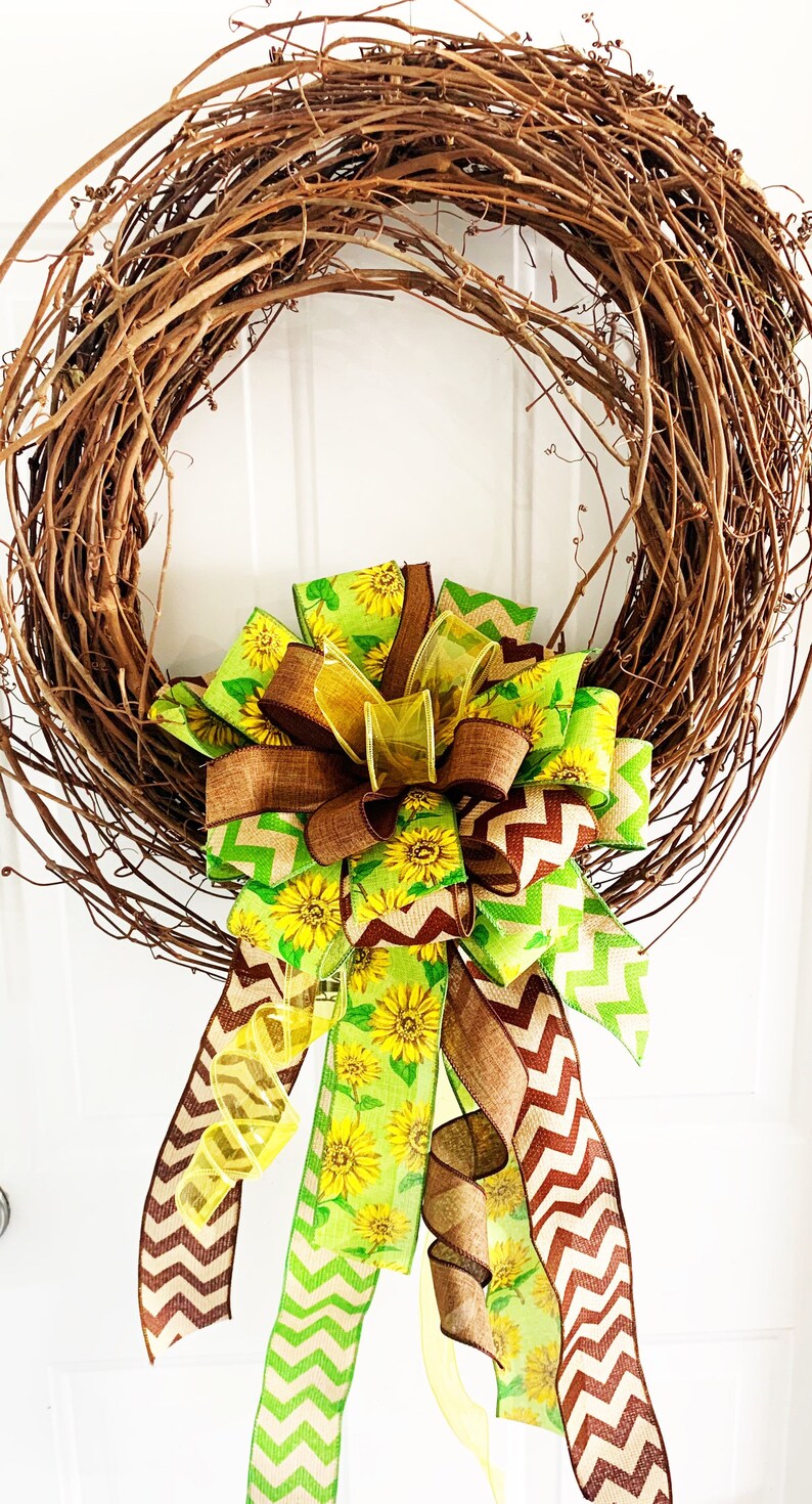 May include: A grapevine wreath adorned with a large bow. The bow features ribbons with sunflower patterns, brown chevron stripes, and solid brown and yellow accents. The wreath is a decorative item for doors or walls.
