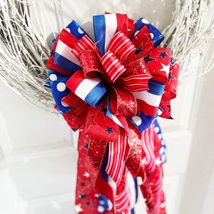 May include: A patriotic wreath bow featuring red, white, and blue ribbons. The bow has a layered design with various patterns, including stars, stripes, and polka dots. The bow is attached to a white twig wreath, perfect for Independence Day or other patriotic holidays.