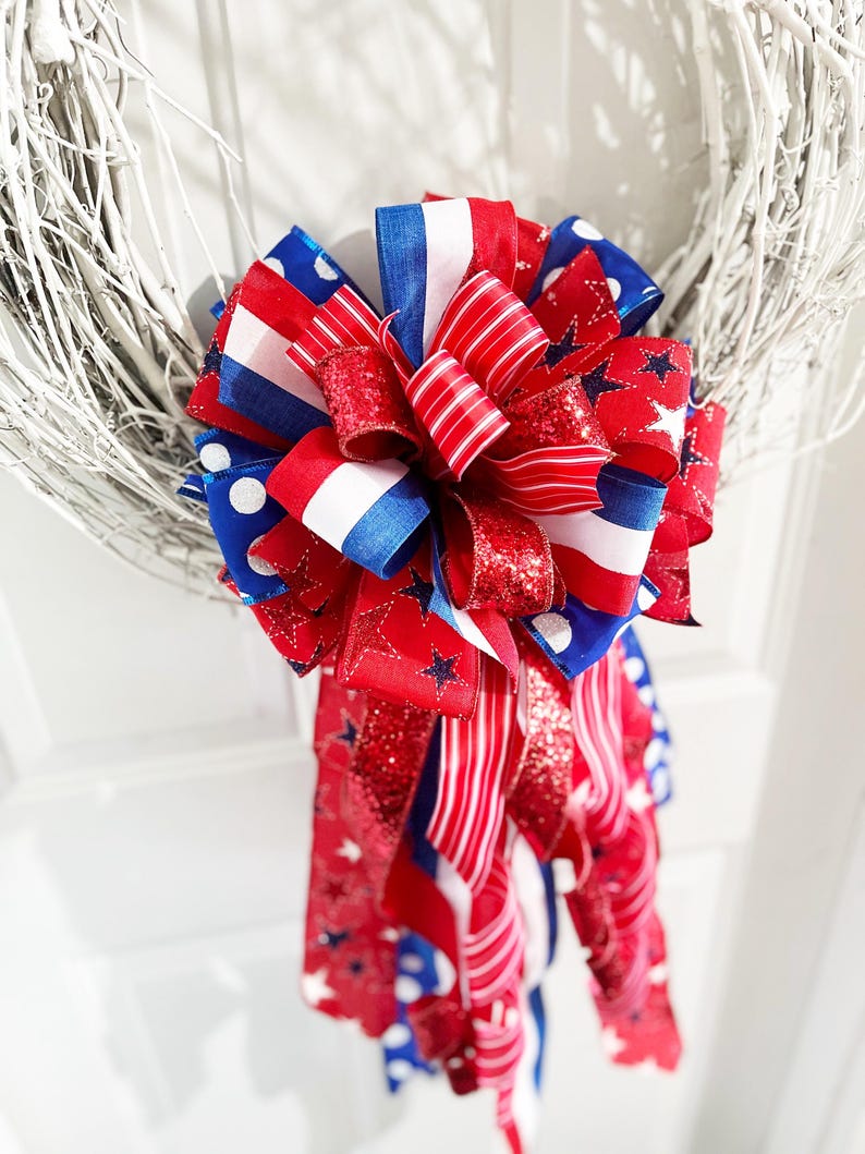 May include: A patriotic wreath bow featuring red, white, and blue ribbons. The bow has a layered design with various patterns, including stars, stripes, and polka dots. The bow is attached to a white twig wreath, perfect for Independence Day or other patriotic holidays.
