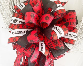 May include: A red and black bow with paw prints and the text "Georgia" on it. The bow is tied with a plaid ribbon.