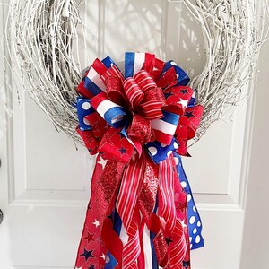 May include: A white twig wreath adorned with a large bow featuring red, white, and blue ribbons. The bow includes ribbons with stars, stripes, and polka dots, creating a patriotic theme. Perfect for Independence Day or other patriotic holidays.