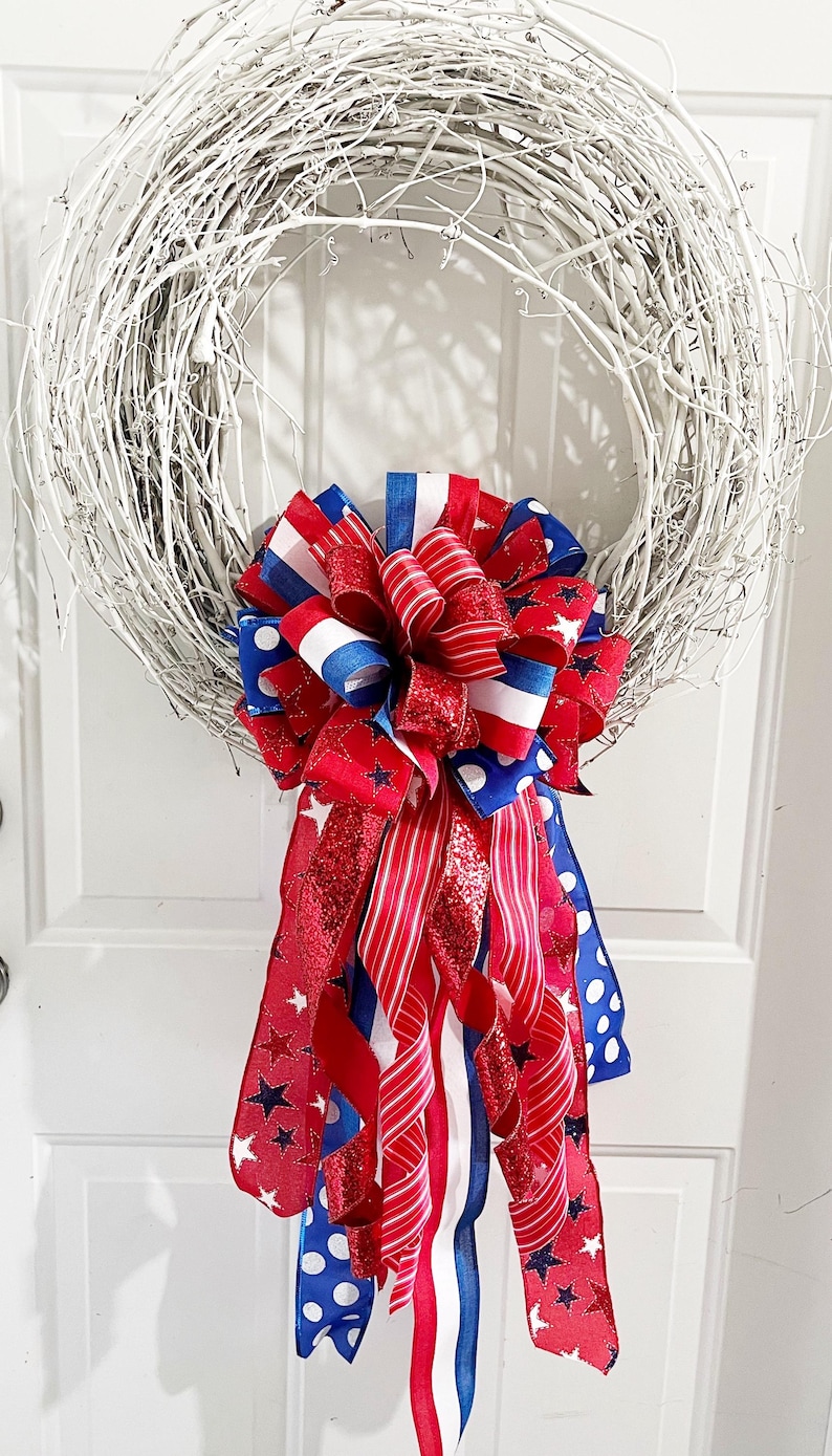 May include: A white twig wreath adorned with a large bow featuring red, white, and blue ribbons. The bow includes ribbons with stars, stripes, and polka dots, creating a patriotic theme. Perfect for Independence Day or other patriotic holidays.